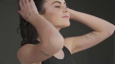 Siyah eşofmanlı, güzel, gülümseyen genç bir kadın sabah antrenmanına gri bir stüdyonun penceresinin yanında başlıyor.