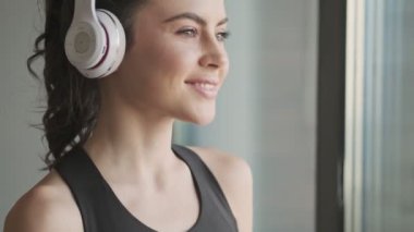 Siyah eşofmanlı mutlu bir genç kadın gri bir stüdyonun penceresinin yanında duran kulaklıkla müzik dinliyor.