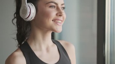 Siyah eşofmanlı hoş bir genç kadın gri bir stüdyonun penceresinin yanında duran kulaklıkla müzik dinliyor.