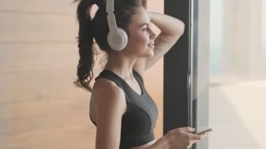 Siyah eşofmanlı mutlu genç bir kadının yan görüntüsü, gri bir stüdyonun penceresinin yanında dururken akıllı telefonuyla müzik dinliyor.