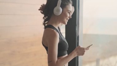 Siyah eşofmanlı çekici bir genç kadının yan görüntüsü, gri bir stüdyonun penceresinin yanında dururken akıllı telefonuyla müzik dinliyor.
