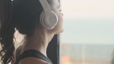 Siyah eşofmanlı güzel genç bir kadın gri bir stüdyonun penceresinin yanında dururken kulaklıkla müzik dinliyor.