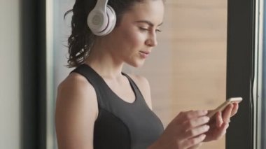 Siyah eşofmanlı, kulaklıklı, sevimli bir genç kadın gri bir stüdyonun penceresinin yanında durup akıllı telefonundan gelen müziği dinliyor ve ona bakıyor.