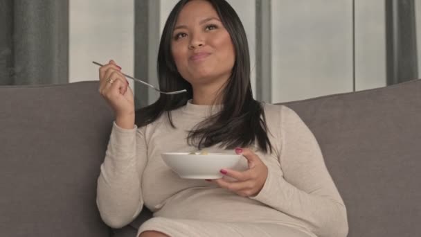 幸せなリラックスした若いです妊娠中のアジアの女性ありますPattating彼女の腹ながら食べますフルーツサラダと見ますテレビで自宅でリビングルーム — ストック動画