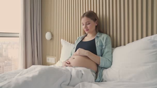 幸せな妊娠中の女性笑顔と彼女の腹をペッティングしながら 日当たりの良い部屋にベッドに横たわって — ストック動画