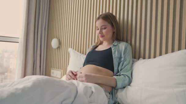 日当たりの良い部屋にベッドの上に横たわっている間 彼女の腹を撫でて笑顔妊娠中の女性 — ストック動画