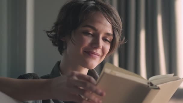 快乐的年轻漂亮的女人坐在沙发上看书 — 图库视频影像