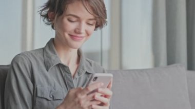 Evdeki kanepede otururken akıllı telefondan mesaj yazan mutlu genç kadın.