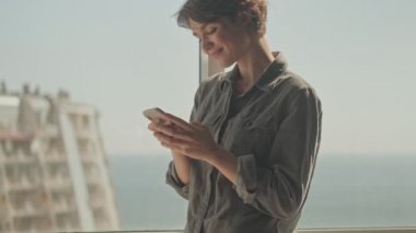 Gülümseyen genç ve güzel bir kadın, akıllı telefon kullanarak evin penceresinin yanında dikiliyor.