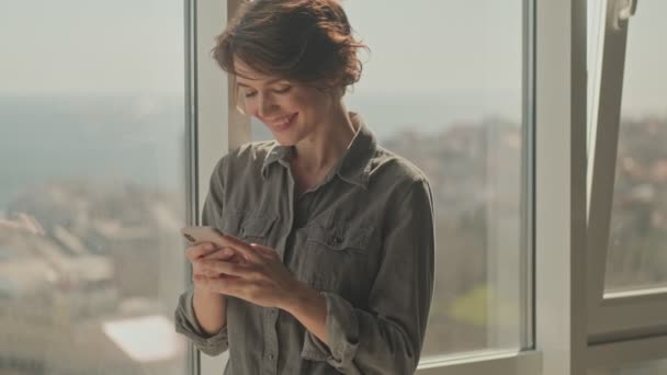Jovem Mulher Bonita Feliz Usando Smartphone Enquanto Está Perto Janela — Vídeo de Stock