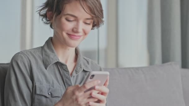 Evdeki Kanepede Otururken Akıllı Telefondan Mesaj Yazan Mutlu Genç Kadın — Stok video