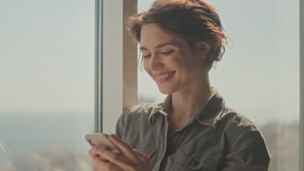 Zadowolony Młoda Ładna Kobieta Pomocą Smartfona Stojąc Pobliżu Okna Domu — Wideo stockowe