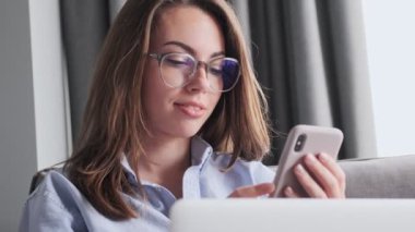 Gözlüklü çekici esmer kadın evinde kanepede otururken akıllı telefon ve dizüstü bilgisayar kullanıyor.