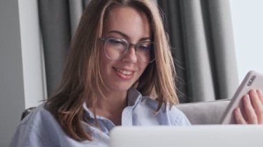 Gözlüklü çekici esmer kadın evinde kanepede otururken akıllı telefon ve dizüstü bilgisayar kullanıyor.