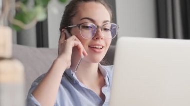 Gözlüklü çekici esmer kadın evinde koltuğun yanında dizüstü bilgisayarla otururken akıllı telefonla konuşuyor.