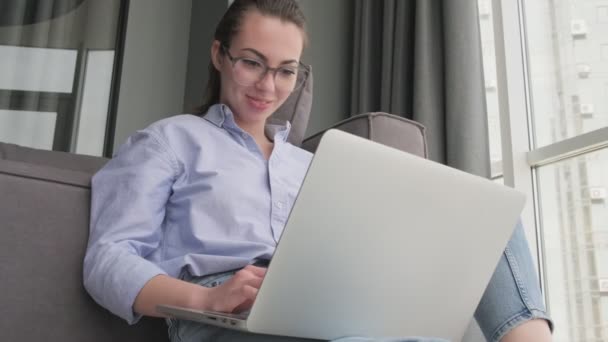 下からの眺め自宅でソファの近くの床に座っている間 ラップトップコンピュータを使用して眼鏡で満足魅力的なブルネットの女性 — ストック動画