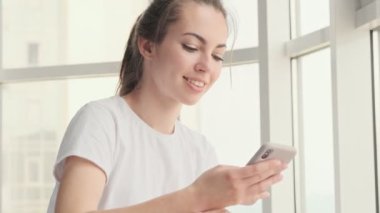 Gülümseyen, esmer, çekici bir kadın. Evinin penceresinin yanında otururken akıllı telefon kullanıyor.