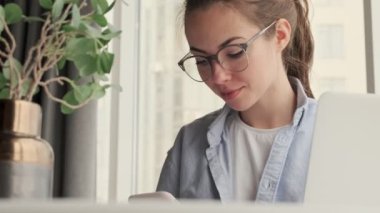 Gözlüklü çekici bir kadın evde dizüstü bilgisayarla çalışırken akıllı telefon kullanıyor.