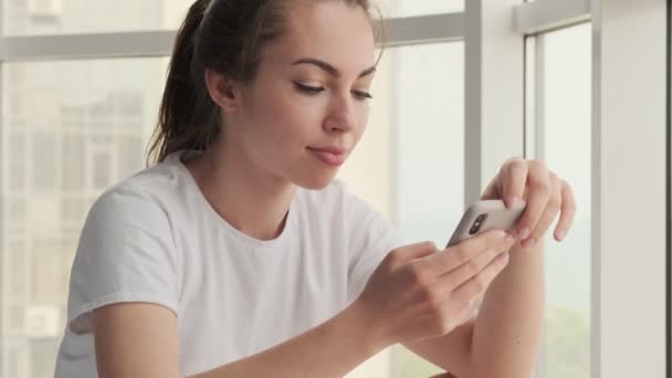 Satisfeito Atraente Mulher Morena Usando Smartphone Enquanto Sentado Perto Janela — Vídeo de Stock