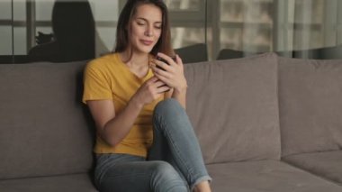 Yakışıklı genç bir kadın evdeki oturma odasındaki kanepede otururken akıllı telefonunu kullanıyor.