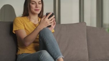 Gülümseyen genç bir kadın evdeki oturma odasındaki kanepede otururken akıllı telefonunu kullanıyor.
