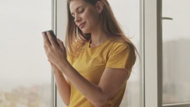 Güzel bir genç kadın akıllı telefonunu kullanıyor. Evinin penceresinin yanındaki oturma odasında duruyor.