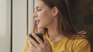 Mükemmel genç bir kadın akıllı telefonunu kullanıyor. Evinin penceresinin yanındaki odada duruyor.