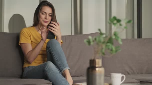 Uma Jovem Satisfeita Está Usando Seu Smartphone Enquanto Senta Sofá — Vídeo de Stock