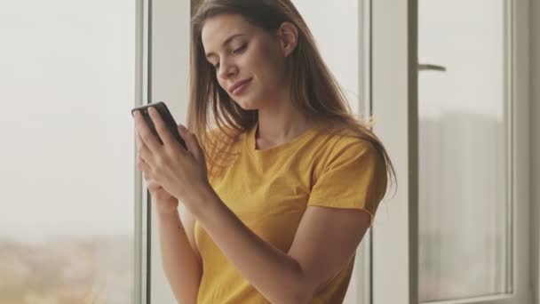 Zadowolona Młoda Kobieta Patrzy Swojego Smartfona Stojącego Salonie Pobliżu Okna — Wideo stockowe
