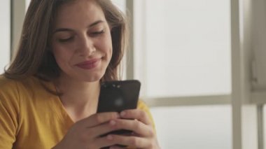 Memnun genç bir kadın evinde akıllı telefonuyla bir şeyler yazıyor.