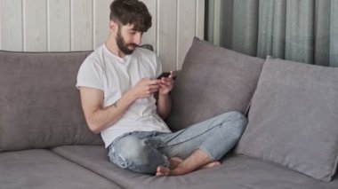 Gündelik giysiler içinde gülümseyen genç bir hippi evinde oturma odasında akıllı telefonunu kullanıyor.