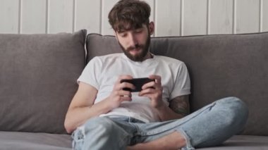 Gündelik giysiler içindeki çekici genç bir hippi evinde oturma odasında oturmuş akıllı telefonuyla bir şey izliyor.