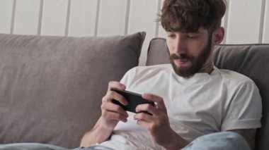 Sıradan giysiler içindeki çekici genç bir hippi evinde oturma odasında oturmuş akıllı telefonuyla oyun oynuyor.