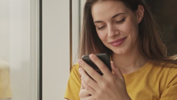 笑顔の若い女性が家の窓の近くの部屋に立っているスマートフォンを使っている — ストック動画