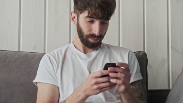 Ein Fokussierter Junger Mann Sitzt Mit Seinem Smartphone Heimischen Wohnzimmer — Stockvideo