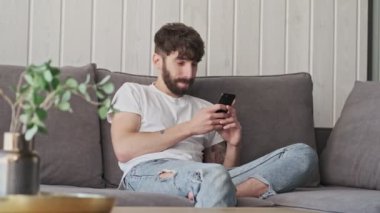 Mutlu gülümseyen, sıradan giyinmiş genç bir hippi akıllı telefonunu evdeki oturma odasında kullanıyor.