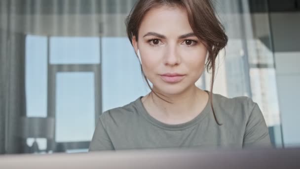 自宅に座っている彼女のラップトップコンピュータを使用している間にイヤホンを持つ魅力的な若い女性が働いています — ストック動画