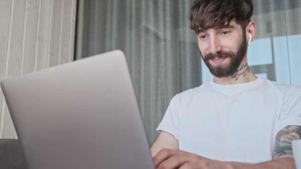 一位穿着休闲装的年轻嬉皮男微笑着坐在客厅里用笔记本电脑 — 图库视频影像