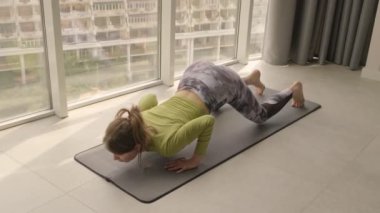 Eşofman giyen genç ve güzel bir kadın sabahları evdeki spor minderinde yoga yapıyor.