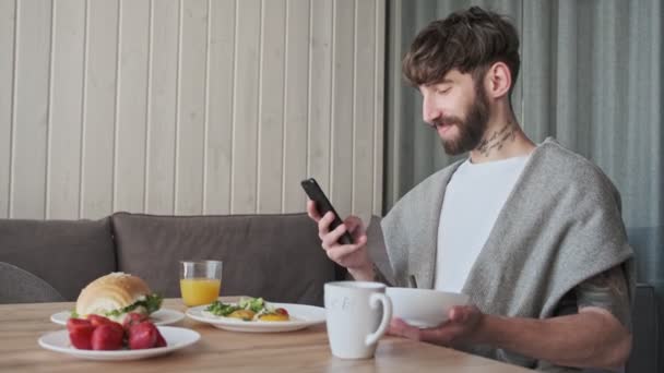 Šťastný Pozitivní Mladý Hipster Muž Používá Svůj Smartphone Při Snídani — Stock video