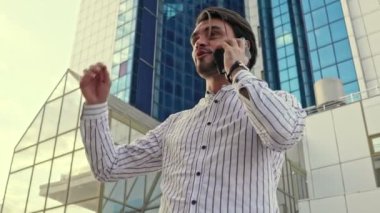 Gözlüklü başarılı bir genç adam modern binaların yakınındaki bir sokakta yürürken akıllı telefonuyla konuşuyor.