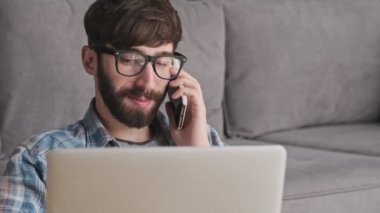 Gözlüklü yakışıklı genç bir hippi, evdeki oturma odasında telefonla konuşurken dizüstü bilgisayarını kullanıyor.