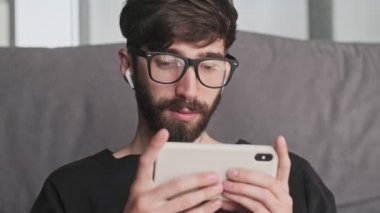 Sakin, genç, gözlüklü bir hippi evinde oturan kulaklıklı akıllı telefonunu kullanıyor.