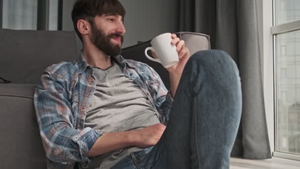 一位穿着休闲装的年轻貌美的嬉皮士男子正坐在客厅里喝着杯子里的东西 — 图库视频影像