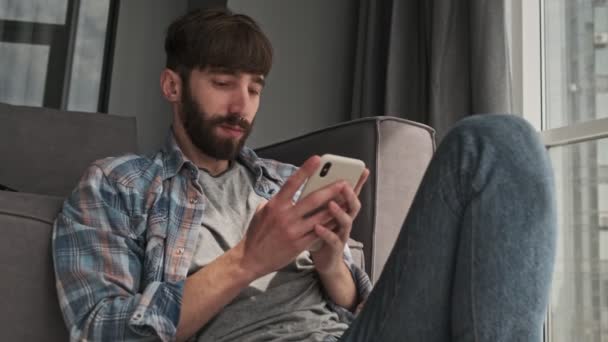カジュアルな服を着た若いヒップスター男性が自宅のリビングルームに座っているスマートフォンを使用しています — ストック動画