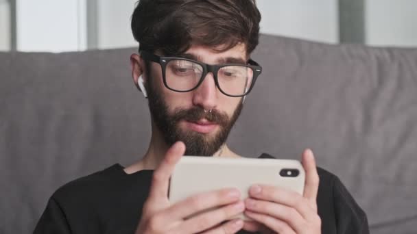 カジュアルな服を着た穏やかな若いヒップスター男性が自宅のリビングルームにイヤホンを装着してスマートフォンを使用しています — ストック動画