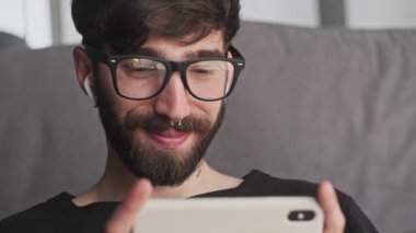 Gözlüklü mutlu genç bir hipster 'ın yakın plan görüntüsü evde oturma odasında oturan kulaklıklı akıllı telefonunu kullanıyor.