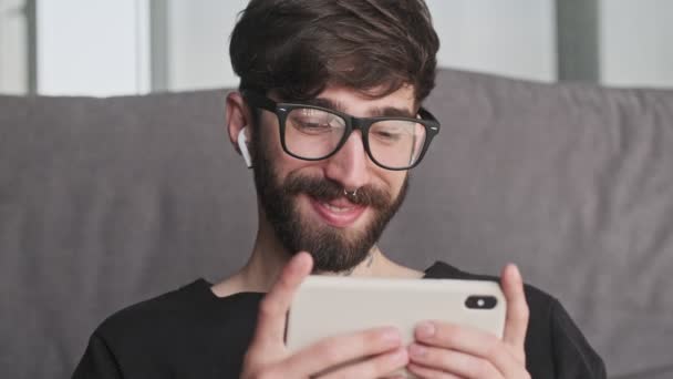 Usmívající Mladý Hipster Muž Brýlemi Používá Svůj Smartphone Sluchátky Sedí — Stock video