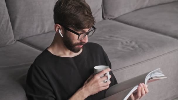 Atractivo Joven Hipster Con Auriculares Está Leyendo Libro Mientras Bebe — Vídeo de stock