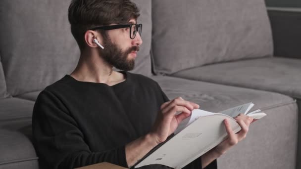 一位年轻迷人的嬉皮士 戴着眼镜 头戴耳塞 坐在客厅里看书 — 图库视频影像